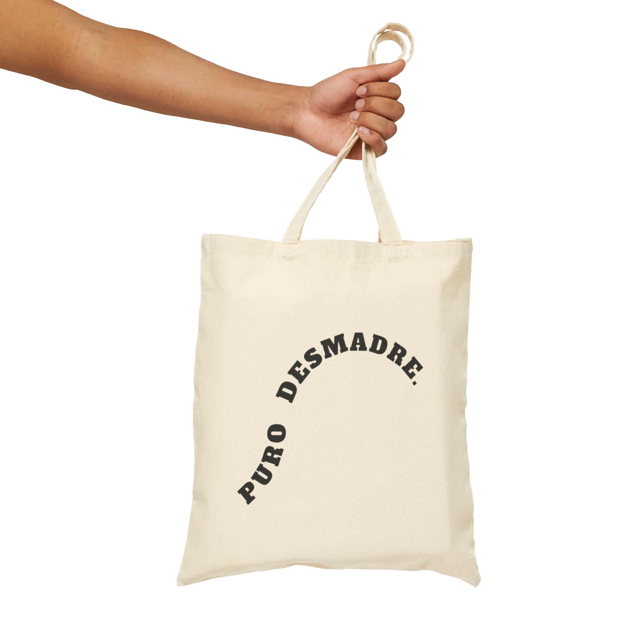 Beige tote bag with 'Puro Desmadre' text held by a hand on a white background