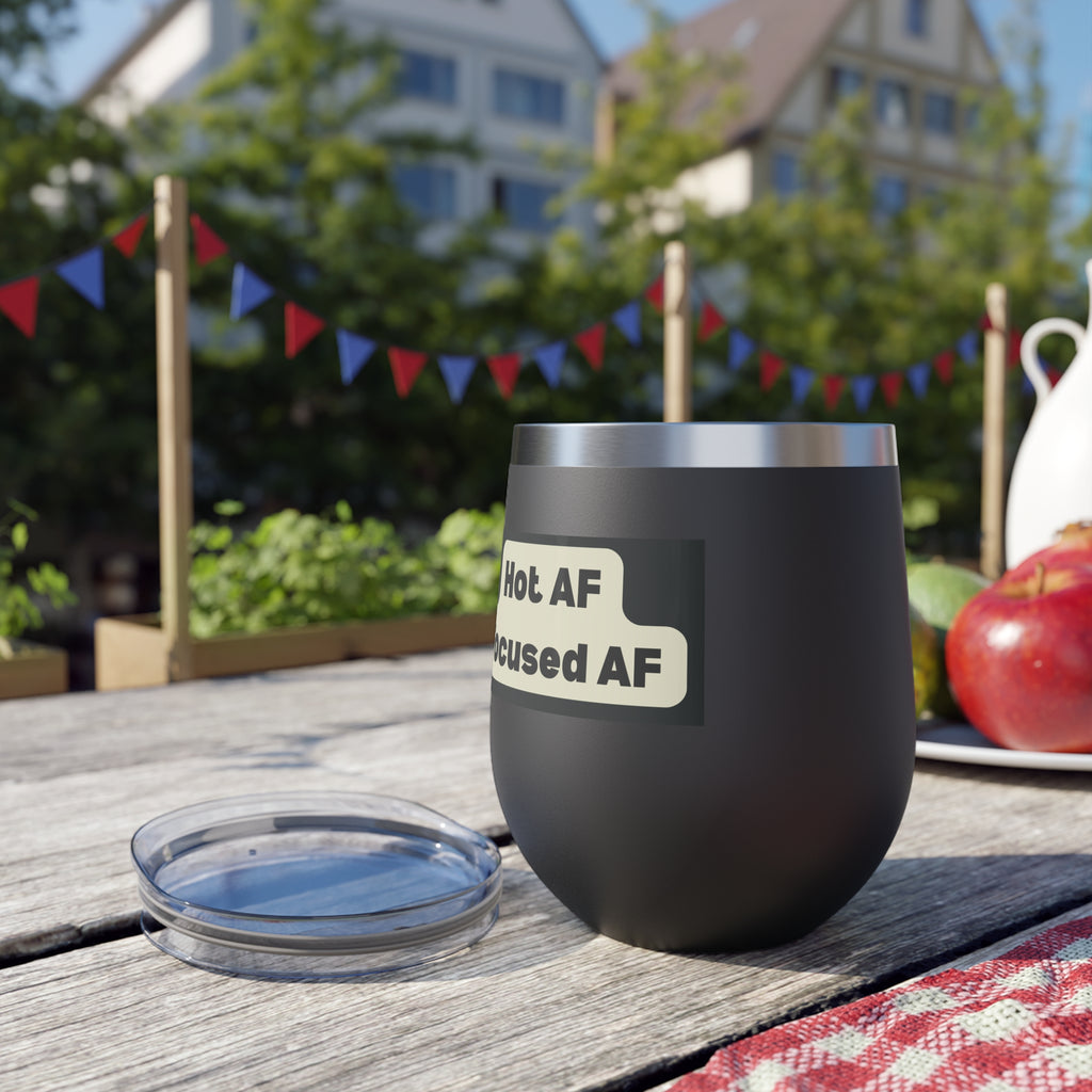 Black insulated wine tumbler with humorous label on a wooden table outdoors.