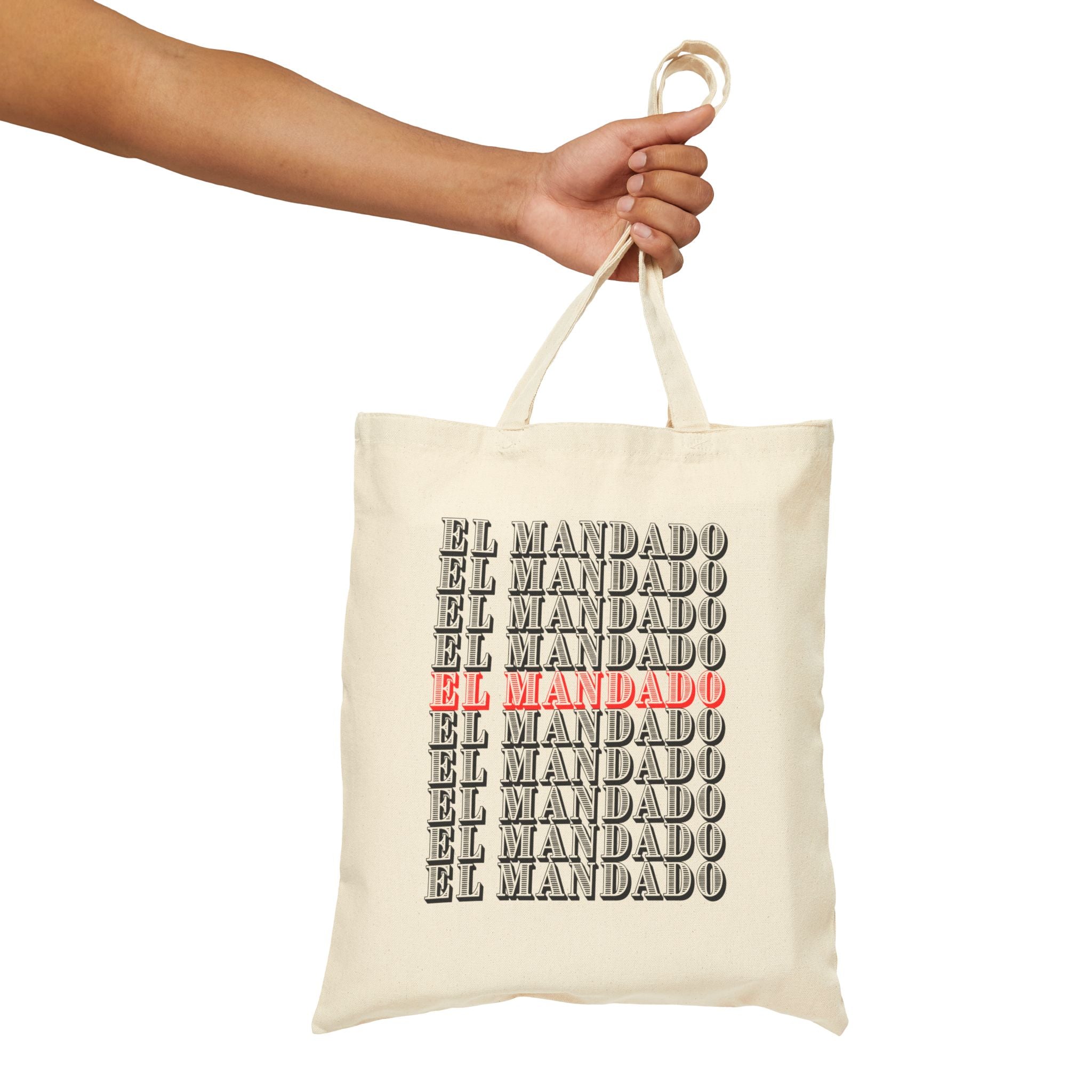 Beige tote bag with 'EL MANDADO' text pattern held by a hand on a white background